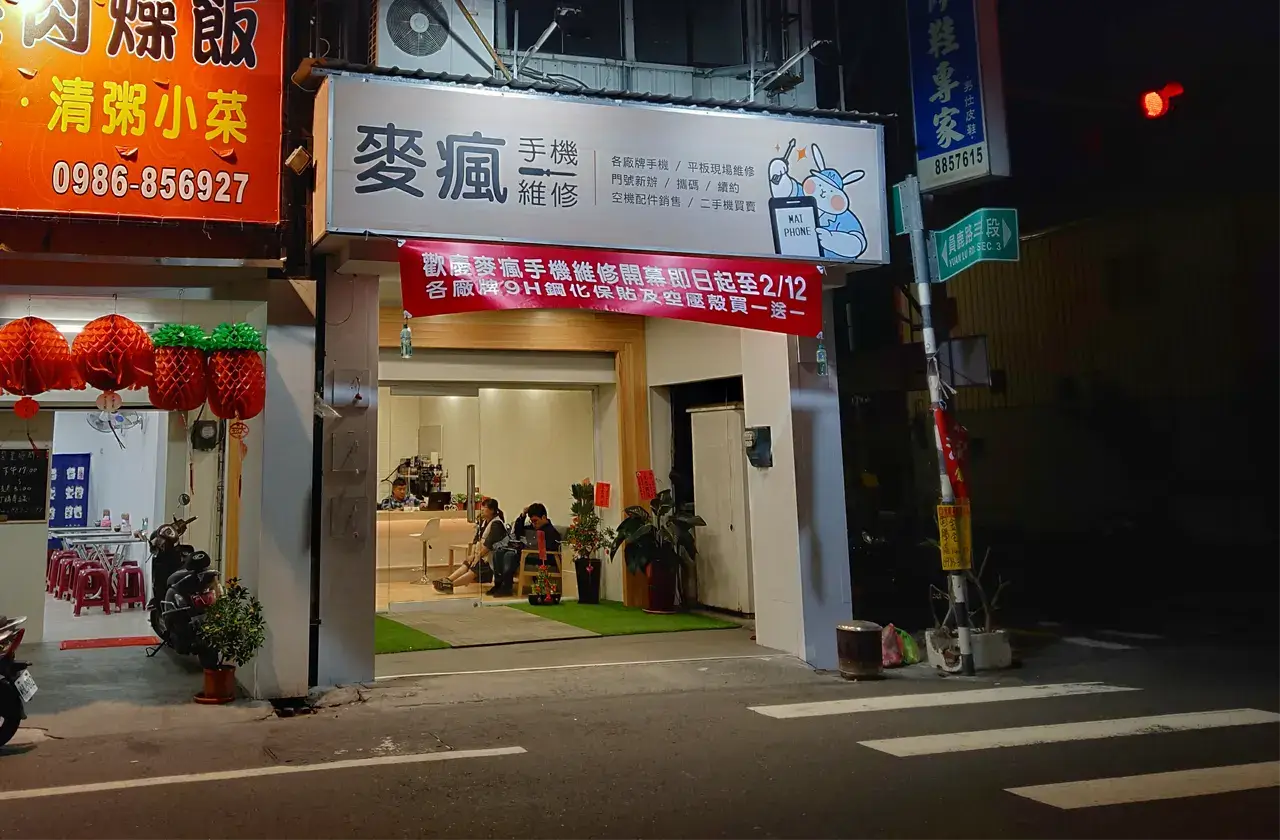 黑夜中的麥瘋手機維修店面門口，明亮招牌與騎樓燈光，照亮路面